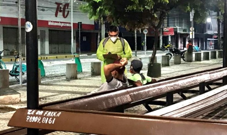 um profissional da prefeitura, com uma prancheta na mão, fala com dois homens sentados em um banco na praça do ferreira à noite